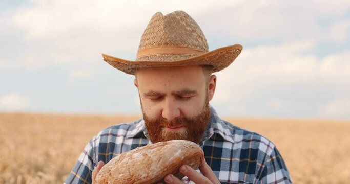 Close up of handsome red-haired male farmer smelling fresh bread while standing in golden field. Portrait of happy countryman in hat with bake standing in rural area in good mood. Agricultor concept