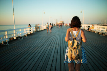 Young female tourist with vintage backpack walking on the wooden pier in golden hour - obrazy, fototapety, plakaty