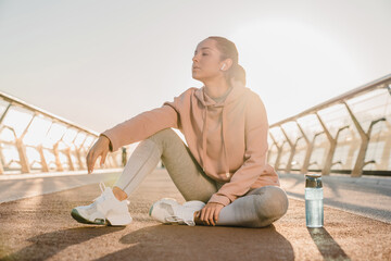 Athletic girl in sports outfit after jogging, sitting on the parapet of the bridge, against the backdrop of white sky with rising sun.