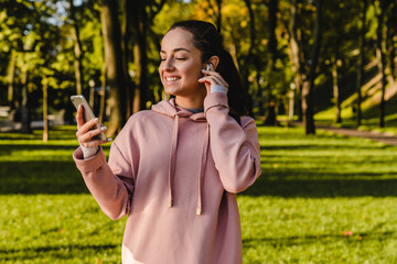 Athletic woman 20s in sportswear in green park using mobile phone with bluetooth earpod