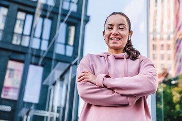 Happy smiling young lady in fitness outfit is standing against urban background