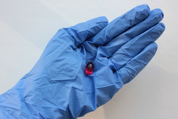 Hand in blue rubber glove with red capsule on white background. Doctor holding pill in hand. 