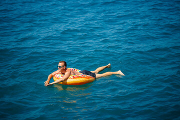 A young man floats on an inflatable air ring circle in the sea with blue water. Festive holiday on a happy sunny day. Vacation concept, top view.