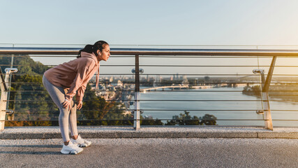 Young lady runner is resting after jogging on the bridge against city landscape