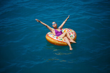 A young beautiful girl in a bright swimsuit lies on a large inflatable ring and floats on the blue sea on a bright sunny summer day