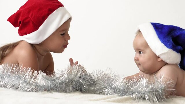 merry xmas and happy new year, infant, childhood, holidays concept - close-up 6 month old newborn naked baby in red and blue santa claus hat with elder sister crawls with decorations christmas tree