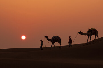 Fototapeta na wymiar Man and a camel walking across sand dunes in Jaisalmer, Rajasthan, India.