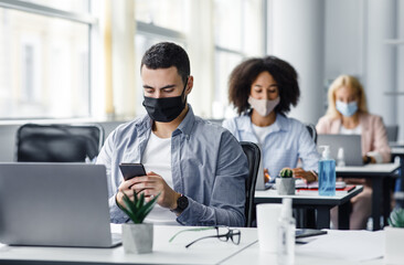 Modern gadgets for working with clients remotely from office after quarantine. Arabic young guy in protective mask typing on smartphone at table with laptop