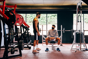 two men wearing face masks talk while working out at the gym