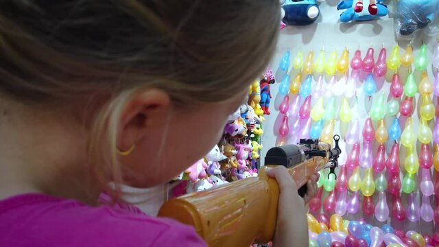 Young Caucasian girl shooting rubber bullet gun at rows of colorful balloons to win prizes at shooting range