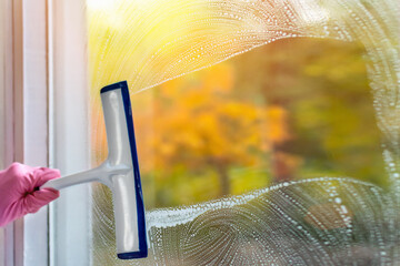Home routine, housework concept. A woman clean a window pane with a squeegee and soap suds. Hands in pink protective gloves cleaning glass on the windows with a detergent spray bottle, copy space.