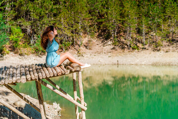 Fototapeta na wymiar Young woman stands on a wooden bridge with raised arms up on the nature background with an emerald mountain lake. Travel, Freedom, Lifestyle concept.