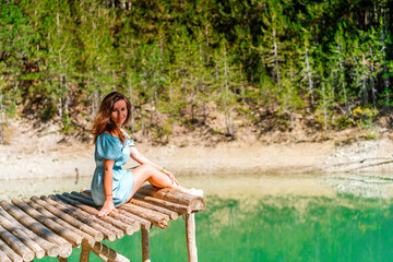 Naklejka na ściany i meble Beautiful girl with long hair in a dress on a bridge above a mountain lake with clear water and a view of a green forest. Copy space