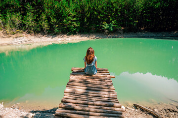 Rear view of a female traveler sitting on a wooden bridge and look at the mountain lake. Travel and active life concept. Adventure and travel in the mountain