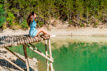 Fototapeta na wymiar Young woman stands on a wooden bridge with raised arms up on the nature background with an emerald mountain lake. Travel, Freedom, Lifestyle concept.