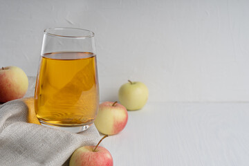 Organic vegetarian apple juice in drinking glass standing on textile towel on white wooden background surrounded by raw ripe fruits showing healthy lifestyle and vegan eating. Image with copy space