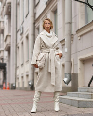 Elegant woman in white dress, hessian boots and coat walking at city street. Fall autumn fashion look. Pretty tall stylish young gitl with fashionable makeup and hair style. Elegant lady. Full length