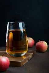 Organic tasty apple juice in drinking glass standing on cutting board on dark wooden background surrounded by raw ripe fruits showing healthy lifestyle and vegetarian eating. Vertical orientation