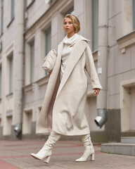 Elegant woman in white dress, hessian boots and coat waling at city street. Fall autumn fashion look. Pretty tall stylish young gitl with fashionable makeup and hair style. Elegant lady. Full length