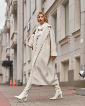 Elegant woman in white dress, hessian boots and coat walking at city street. Fall autumn fashion look. Pretty tall stylish young gitl with fashionable makeup and hair style. Elegant lady. Full length