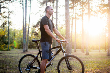 A caucasian man in a black t-shirt and shorts is staying with a black bike in a forest in the...