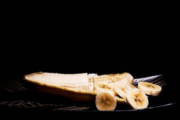 Plátano semi pelado con la mitad en rodajas sobre plato negro de madera labrada.