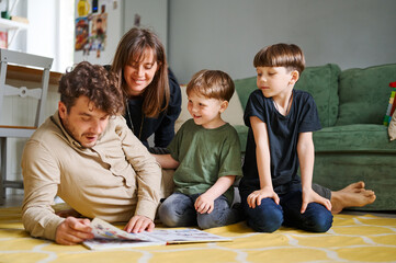 Fototapeta premium Happy family with two little sons reading story indoors, parents with children spending time together and lying on the floor at home