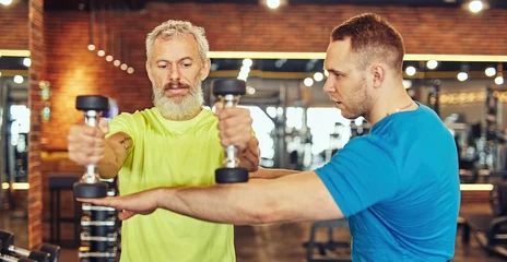 Foto op Canvas Focused mature caucasian man in sportswear doing weight exercises with assistance of a young professional personal trainer © Kostiantyn