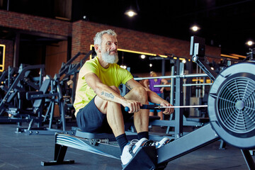 Hard workout. Mature athletic man in sportswear exercising on rowing machine at gym