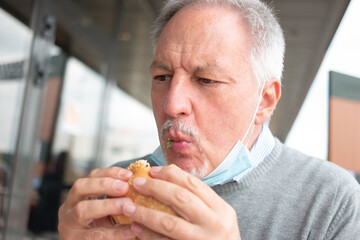 Man eating his fast food lunch hamburger during with a mask on his chin
