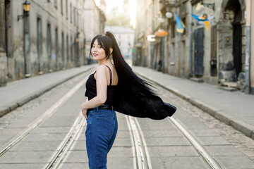 Fototapeta premium Young beautiful Asian girl with ponytail black hair, wearing casual jeans and black top, turning to camera and posing while walking in the city on a sunny day. Outdoor fashion portrait