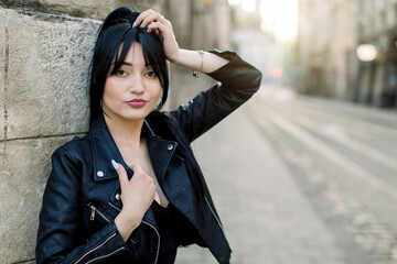 Fototapeta premium Happy mixed raced Caucasian woman in black leather jacket, smiling while posing outdoors in the city at sunny day, leaning on old stone building wall. Outdoor shot, copy space