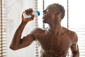 Positive african man singing taking shower holding shampoo bottle indoor