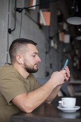 Young man in casual clothes listening to music in wireless earphones while using smartphone in cafe.