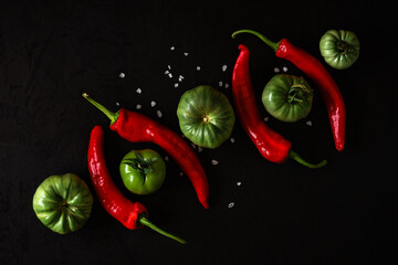artistic combination of alternating chili peppers and green tomatoes with coarse sea salt on a black plastered surface arranged diagonally. view from above. creative dark still life