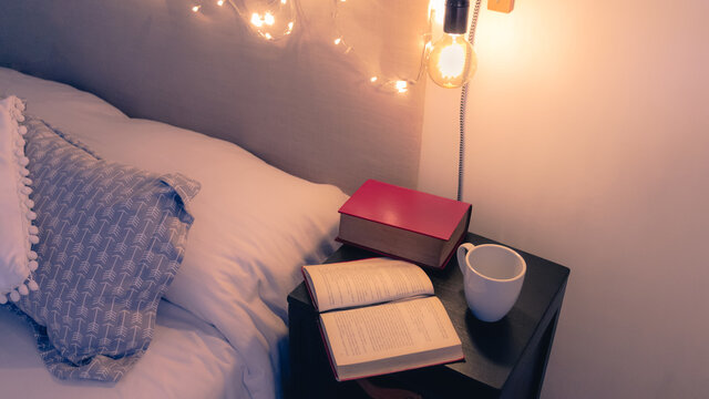 Leer un buen libro antes de dormir. Plano levemente elevado de mesa de noche junto a una cama con libros antiguos y una taza de caf&eacute; en un c&aacute;lida confortable e iluminada habitaci&oacute;n. 