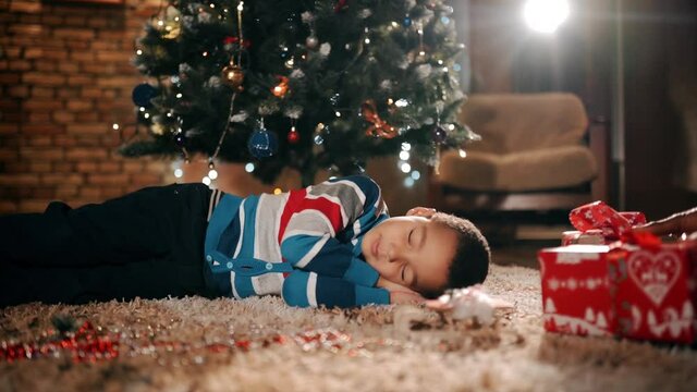 A small child is sleeping next to christmas tree. Christmas presents appear secretly.