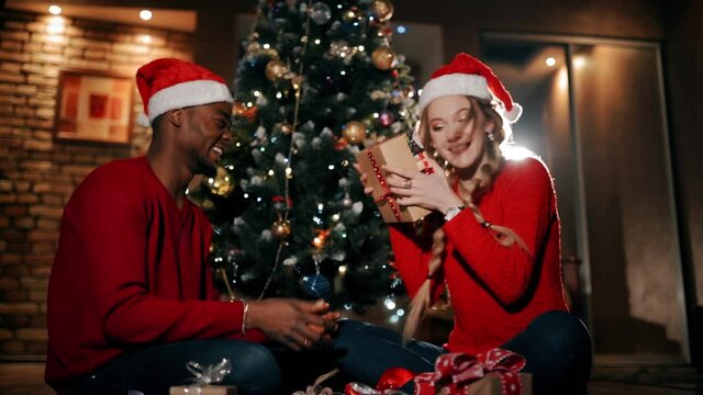 happy couple are sitting in a fluffy carpet in the living room at home. A man is giving a christmas present to girl.