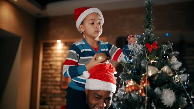 A child is decorating a christmas tree with glittering balls, sitting on his father's shoulders.
