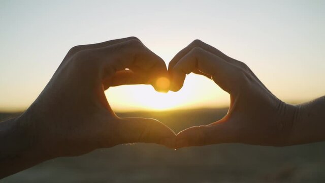 Man and woman hands making heart or love gesture together at desert sunset. Loving couple doing romantic gesture with hands - heart symbol with sun rising or setting in background. Close up.