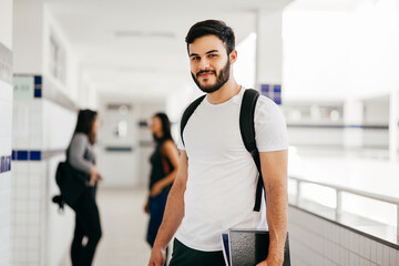 Portrait of young Brazilian student with backpack carrying books in college. Other students in the background. - obrazy, fototapety, plakaty