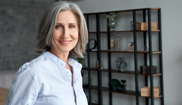 Smiling confident stylish mature middle aged woman standing at home office. Old senior businesswoman, 60s grey-haired elegant lady executive business leader manager looking at camera, portrait.