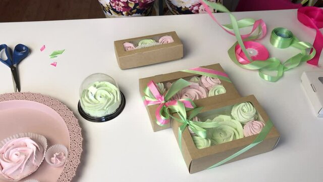 A woman packs pink and light green marshmallows in gift cardboard boxes. Ties them up with a ribbon.