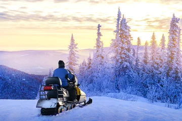 Fotobehang Man standing on snowy mountain near snowmobile enjoying view winter forest at sunset. Concept winter travel, sports, activity. © Sergey