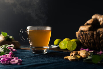 Immune boosting ginger and lemon tea with steam coming out of the cup and a basket full of ginger roots, lime, peppermint leaves and pink flowers. Black background with copy space.