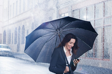 Smiling businesswoman with umbrella and smart phone on rainy street © Martin Barraud/Caia Image