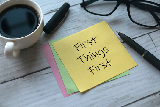 Selective focus of a cup of coffee, pen, glasses and memo notes written with First Things First on white wooden background.