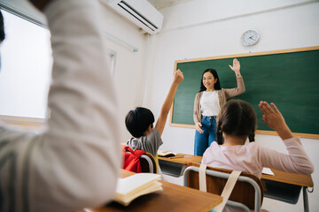 Asian school teacher with students raising hands. Young woman working in school with arm raised, school children putting their hands up to answer question, enthusiasm, eager, enjoyment.