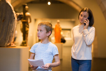 Attentive boy with paper guide looking at exposition in historical museum, woman using phone behind