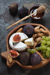 Close-up of a wooden serving tray with camembert cheese, fig fruits, grapes, nuts and honey, vertical shot on a brown stone surface